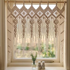 Peut inclure: Un rideau de fenêtre en macramé beige avec un motif géométrique en losanges. Le rideau est suspendu à une tringle en bois, et le design comprend des détails noués et de longues franges. La fenêtre est encadrée par un cadre blanc.