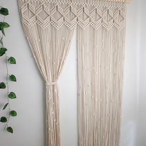 Op de afbeelding: Crèmekleurig macramé gordijn met een geometrisch ontwerp aan de bovenkant. Het gordijn hangt aan een houten roede en is aan één kant vastgebonden. Het macramé heeft een ruitpatroon en kralenaccenten.
