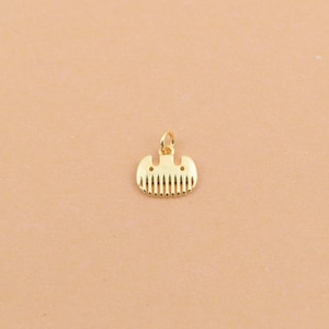Puede incluir: Un pequeño colgante dorado con forma de peine. El colgante tiene una parte superior redondeada con un pequeño lazo para una cadena y una serie de líneas verticales que representan los dientes del peine. El fondo es de color beige claro.