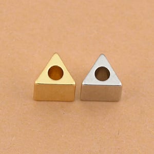 May include: Two small, triangular metal beads. One is gold-colored, the other is silver-colored. Each bead has a circular hole in the center. The beads are likely for jewelry making or other craft projects.