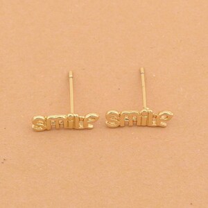 May include: A pair of gold-colored stud earrings. Each earring spells out the word "smile" in a delicate, cursive font. The earrings are set against a neutral background.