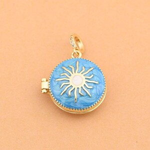 May include: A gold pendant with a light blue enamel design featuring a white sunburst. The round pendant has a hinged opening and a small loop for a chain. The pendant is set against a peach-coloured background.