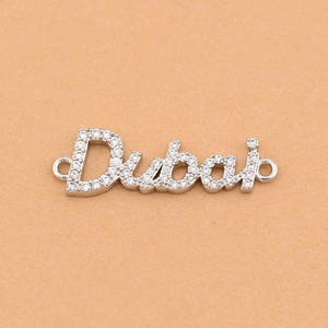 Puede incluir: Un charm plateado con la palabra "Dubai" escrita en cursiva. Las letras están incrustadas con pequeñas piedras preciosas transparentes. El charm tiene pequeños bucles en cada extremo para sujetar.