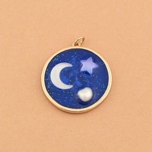 May include: A round pendant with a gold-colored border. The pendant features a dark blue background with a white crescent moon, a purple star, and a white pearl. The design is reminiscent of a night sky.