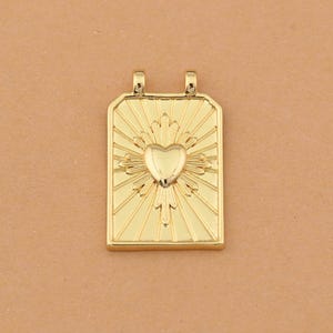 以下が含まれることがあります： 中央にハートがあり、太陽光線が放射状に広がる金色のペンダント。ペンダントは長方形で、チェーンを通すための小さなループが上部に2つあります。背景は無地のタン色です。