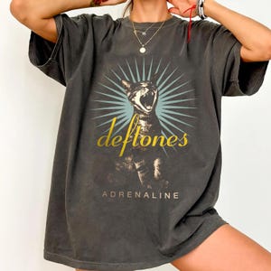 May include: Dark gray t-shirt featuring a graphic of a cat with an open mouth, set against a blue burst design. The word "Deftones" is in gold script above the cat, with "ADRENALINE" below. The shirt has short sleeves.