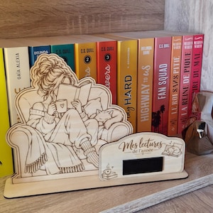 Op de afbeelding: Een lichtgekleurd houten displaystuk met een gegraveerde illustratie van een persoon die in een fauteuil zit en een boek leest met een kat, naast een kleiner bordje met de tekst "Mes lectures de l'année" en een klein zwart rechthoekig inzetstuk.