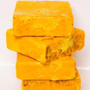 May include: Four stacked, rectangular bars of handmade soap. The soap is a vibrant yellow-orange color with a slightly foamy texture and small, dark specks throughout. The bars are neatly arranged, suggesting a focus on natural skincare.