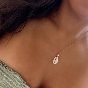 Puede incluir: Un collar de cadena dorada con un colgante de concha blanca. El collar lo lleva una persona con el pelo largo y castaño.