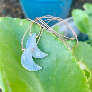 May include: A pair of crescent moon shaped pendants made of white and blue iridescent glass. The pendants are strung on a thin brown cord with gold beads.
