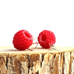 May include: A pair of raspberry-shaped earrings with red, textured surfaces. The earrings are displayed on a small, round, wooden slice with a light brown bark. The background is white.