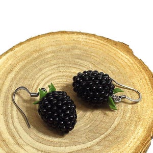 Op de afbeelding: Een paar zwarte braam oorbellen met zilverkleurige haken. De oorbellen zijn gedetailleerd met kleine, ronde kralen om de textuur van de vrucht na te bootsen. De oorbellen worden gepresenteerd op een lichtbruine houten schijf.