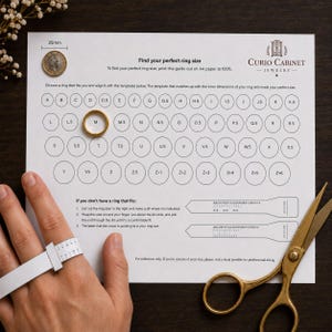 Può includere: Una guida cartacea bianca per la misura degli anelli con una griglia di cerchi etichettati da A a Z+6, un anello dorato sul cerchio M, una moneta da 25mm per la scala, istruzioni, due sagome di carta da ritagliare e forbici dorate. Una mano indossa un misuratore di anelli.