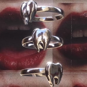 May include: Three silver-toned rings, each featuring a prominent, polished tooth-shaped centrepiece. The rings have a smooth, metallic finish and an open-band design, showcasing a reflective surface that captures light across the tooth motif.