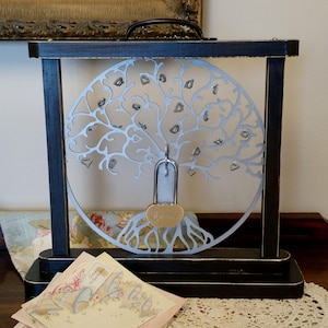 May include: A black wooden frame with a silver metal tree design. The tree has many small hearts hanging from its branches. A padlock with a heart-shaped keyhole is attached to the tree. The padlock has the word "Forever" engraved on it. The frame is sitting on a table with a lace tablecloth.