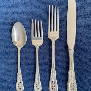 Puede incluir: Juego de cubiertos de plata que incluye una cuchara, dos tenedores y un cuchillo. Los utensilios tienen mangos ornamentados y detallados con un diseño floral. La cuchara y los tenedores tienen tres y cuatro púas, respectivamente. El cuchillo tiene una hoja curva.