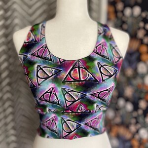 Pode incluir: Um top curto colorido com um padrão repetido de triângulos, círculos e linhas pretas e brancas. O top, exibido em um manequim branco, tem decote redondo. O fundo é uma mistura de tons rosa, verde e azul.