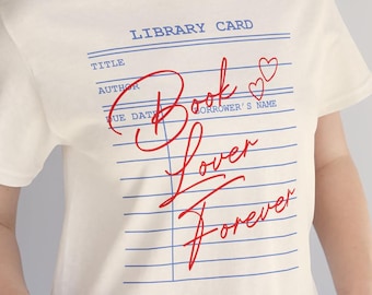May include: A cream-colored short-sleeve t-shirt featuring a blue library card graphic with horizontal lines and the text "LIBRARY CARD," "TITLE," "AUTHOR," and "DUE DATE." Overlaid in red script is the text "Book Lover Forever" with two hearts.