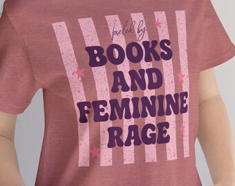 May include: A mauve short-sleeved t-shirt featuring a graphic design with vertical light pink stripes and small pink stars. The text in dark purple reads "fueled by BOOKS AND FEMININE RAGE" in a bold, retro-inspired font style.