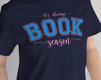 May include: A navy blue short-sleeve t-shirt featuring the text "it's always BOOK season" printed on the front. The word "BOOK" is in large, bold, light blue block letters with a pink outline, while the other words are in a pink script font.