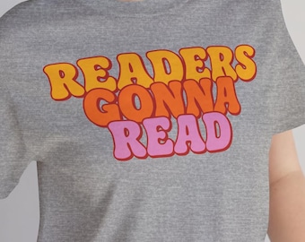 May include: A heather gray short-sleeve t-shirt featuring the text "READERS GONNA READ" in a bold, wavy, retro-style font. The word "READERS" is in yellow, "GONNA" is in orange, and "READ" is in pink, centered on the chest of the shirt.