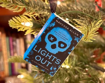 May include: A miniature book ornament with a black ribbon, hanging on a Christmas tree. The book cover is black and blue, with the title "LIGHTS OUT" and a skull graphic. The author's name is "NAVESSA ALLEN".