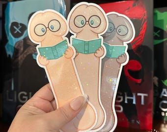 May include: Three phallic-shaped bookmarks with a holographic finish, each featuring a cartoon character wearing round glasses and holding an open teal book. The characters are rendered in shades of light peach, tan, and light brown.
