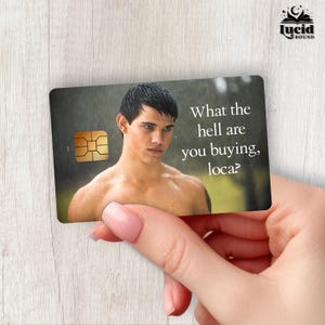 May include: A rectangular credit card skin featuring a shirtless man with dark hair in a rainy setting. The text "What the hell are you buying, loca?" is printed in white on the right side. A gold-colored EMV chip is visible on the left side.
