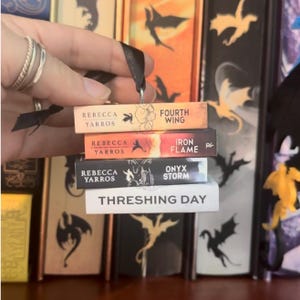 May include: A miniature book ornament stack featuring four rectangular blocks with black ribbon. The spines are labeled Fourth Wing, Iron Flame, Onyx Storm, and Threshing Day, all authored by Rebecca Yarros, with dragon silhouettes and text.