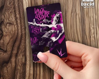 May include: A rectangular card featuring a purple and black graphic of a person singing into a microphone with the text The Vampire Lestat printed in a stylized font. The card has a small notch at the bottom center and a pink logo in the corner.
