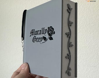 May include: A light gray book with a black ribbon bookmark and a silver charm. The front cover features the black text Morally Grey with a rose illustration. The spine has a vertical vine design with leaves in a dark gray color against a ribbed texture.