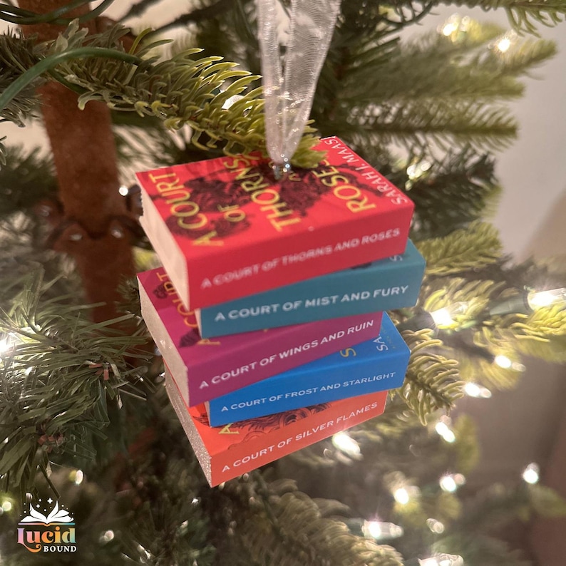 Puede incluir: Un adorno en miniatura con una pila de cinco libros de Sarah J. Maas: A Court of Thorns and Roses, A Court of Mist and Fury, A Court of Wings and Ruin, A Court of Frost and Starlight y A Court of Silver Flames, colgado de una cinta blanca.