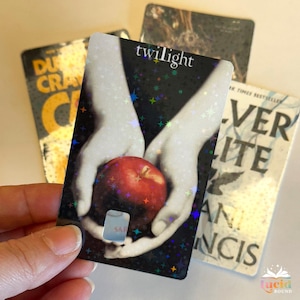 Op de afbeelding: Een holografische boekenlegger met het woord "twilight" en een rode appel in handen. Andere boekomslagen zijn zichtbaar op de achtergrond. De boekenlegger heeft een uitsparing.