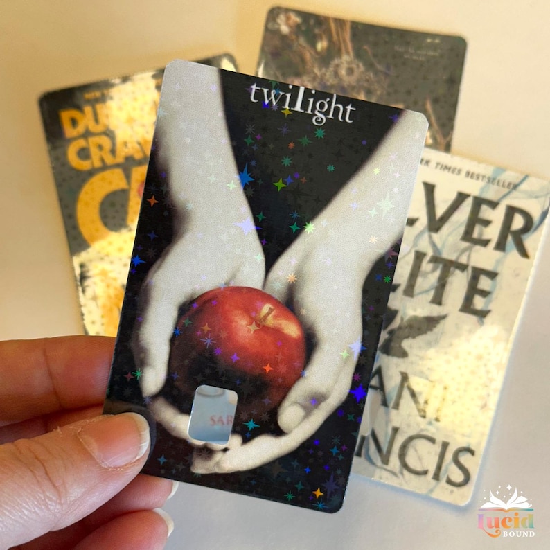 Op de afbeelding: Een holografische boekenlegger met het woord "twilight" en een rode appel in handen. Andere boekomslagen zijn zichtbaar op de achtergrond. De boekenlegger heeft een uitsparing.
