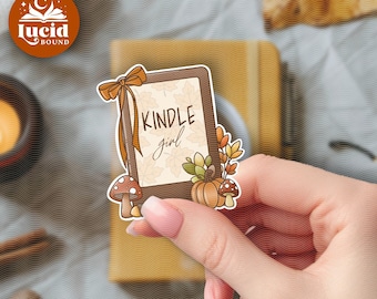 May include: A rectangular sticker featuring a brown frame with a bow at the top left corner. The center displays the text "KINDLE girl" over a light background with leaf patterns. The design includes a pumpkin, mushrooms, and autumn foliage.