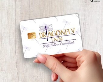 May include: A white rectangular plastic card featuring a purple dragonfly graphic and the gold text Dragonfly Inn with the location Stars Hollow, Connecticut printed below. The card includes a gold metallic chip on the left side and a repeating pattern of light purple dragonfly silhouettes across the background.