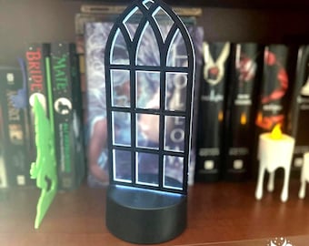 May include: A black, arched window-shaped LED lamp with a grid frame design. The lamp features a solid black circular base and emits a soft, cool-toned light through the window panes, creating a glowing effect against the dark, book-filled background.