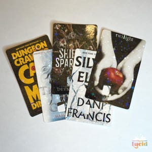 Op de afbeelding: Een verzameling van vijf rechthoekige kaarten met boekomslagen. De titels zijn onder meer "Dungeon Crave", "Ship of Sparks" en "Twilight". De "Twilight"-kaart toont handen die een rode appel vasthouden. De kaarten hebben een glanzend, holografisch effect.