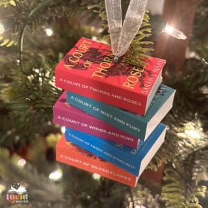 May include: A stack of miniature book ornaments with titles from the "A Court of Thorns and Roses" series by Sarah J. Maas. The books are red, teal, purple, blue, and orange, with white text and a silver ribbon for hanging.