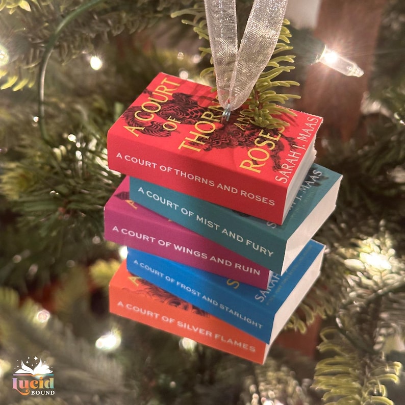 Puede incluir: Un adorno de libros en miniatura que muestra una pila con los t&iacute;tulos A Court of Thorns and Roses, A Court of Mist and Fury, A Court of Wings and Ruin, A Court of Frost and Starlight y A Court of Silver Flames, colgado de una cinta blanca.