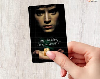 May include: A rectangular credit card featuring a dark, close-up portrait of a person with intense eyes. The text "one cha-ching to rule them all" is printed in white script. A gold-colored EMV chip is positioned near the bottom center.