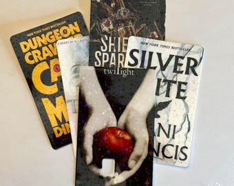 May include: A collection of five rectangular book-themed plastic cards featuring various cover art designs. The cards display titles including Dungeon Crawler Carl, Shield of Spark, and Twilight, with one card showing hands holding a red apple.