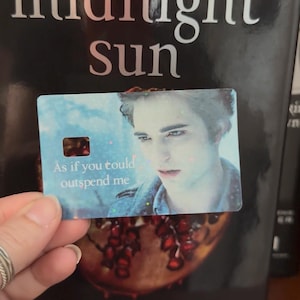 May include: A rectangular card featuring a holographic finish and a portrait of a man with dark hair. The text "As if you could outspend me" is printed in white on the lower left side. The card has a small rectangular cutout on the left side.