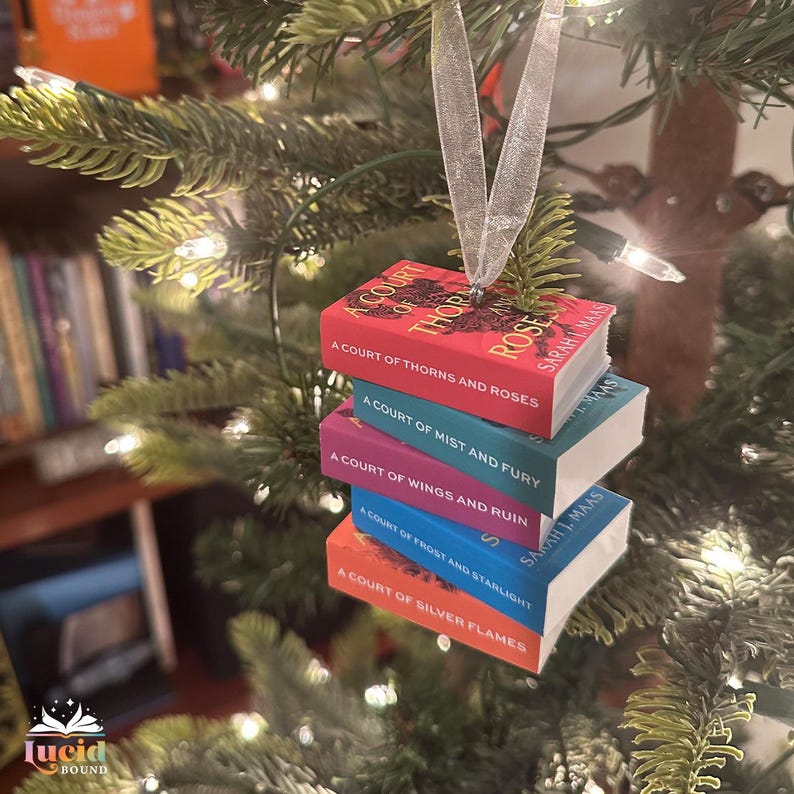 Puede incluir: Un adorno en miniatura de una pila de libros con cinco t&iacute;tulos de Sarah J. Maas: A Court of Thorns and Roses, A Court of Mist and Fury, A Court of Wings and Ruin, A Court of Frost and Starlight y A Court of Silver Flames, suspendido por una cinta plateada.