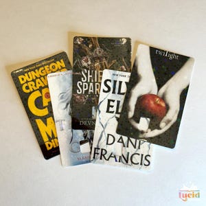 Op de afbeelding: Vijf rechthoekige bladwijzers met boekomslagontwerpen. Titels zijn onder meer "Dungeon Crave", "Ship of Sparks", "Silver El" en "Twilight". De "Twilight"-bladwijzer toont handen die een rode appel vasthouden.