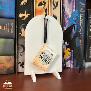 Può includere: Un espositore per libri bianco ad arco con un ornamento a forma di libro in miniatura. La copertina del libro recita "Fourth Wing" di Rebecca Yarros. Un nastro nero sospende il libro. Lo sfondo presenta una libreria con vari libri e decorazioni a tema drago.