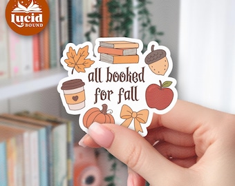 May include: A die-cut sticker featuring the text all booked for fall in dark brown lettering, surrounded by illustrations of a stack of books, an acorn, a red apple, a pumpkin, a coffee cup with a heart, a maple leaf, and a light orange bow.