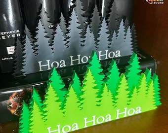 May include: A layered green acrylic forest scene featuring a series of pine trees with the white text "Hoa Hoa Hoa" printed on the front. Behind it sits a similar dark gray acrylic forest scene with the white text "Hoa Hoa Hoa" printed on it.