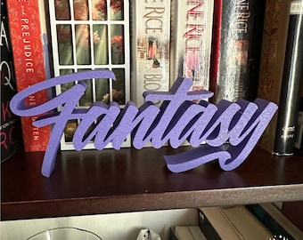 May include: A purple, three-dimensional, freestanding sign featuring the word Fantasy in a cursive script font. The letters have a textured, glittery finish and are positioned on a dark shelf in front of several books and a white decorative frame.