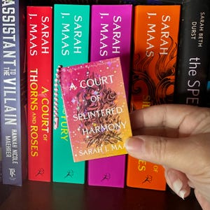 May include: A miniature book keychain titled A Court of Splintered Harmony by Sarah J. Maas, featuring a gradient orange and pink cover with star illustrations, held in front of a row of full-sized books with colorful spines by the same author.